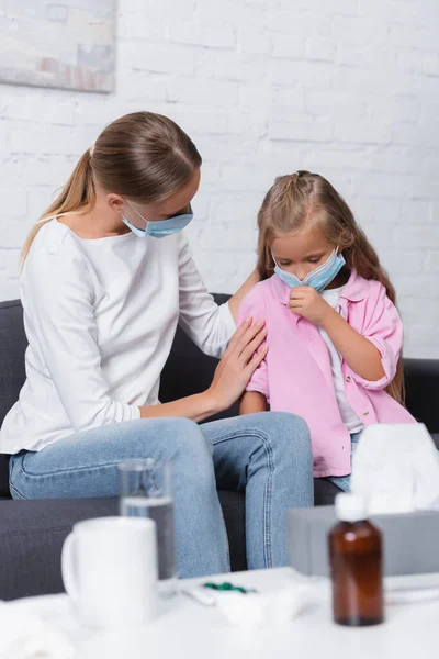 Selective focus of woman in medical mask hugging ill daughter near syrup and napkins on coffee table — Stock Photo