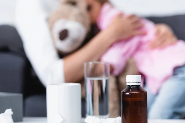 Enfoque selectivo de la botella de jarabe, taza y vaso de agua cerca de madre abrazando niño - foto de stock