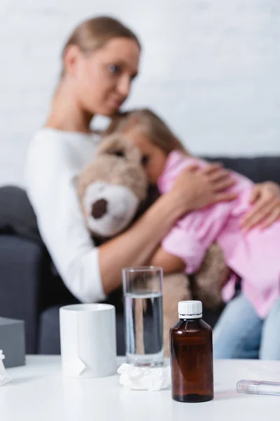 Focus selettivo di sciroppo, bicchiere d'acqua e tazza vicino alla madre che abbraccia il bambino a casa — Foto stock