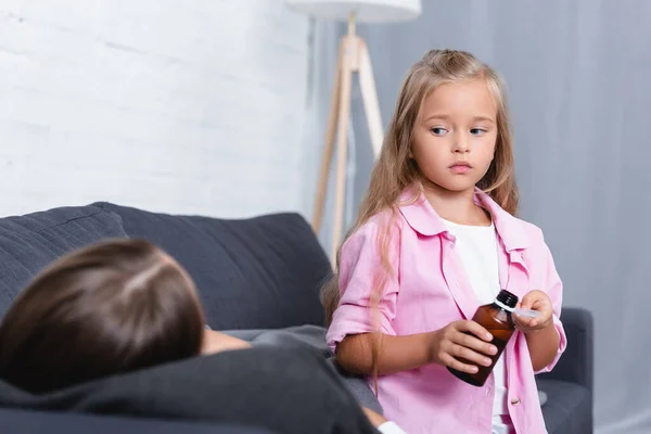 Enfoque selectivo del niño sosteniendo cuchara y jarabe cerca de la madre enferma en el sofá - foto de stock