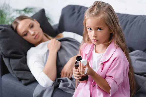 Enfoque selectivo de la niña mirando a la cámara mientras sostiene el jarabe y la cuchara cerca de la madre - foto de stock