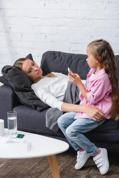 Selective focus of sick woman embracing kid with thermometer near pills and water on coffee table — Stock Photo