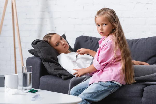 Selektiver Fokus des Mädchens mit Thermometer in der Nähe der kranken Mutter und Tabletten auf dem Couchtisch — Stockfoto