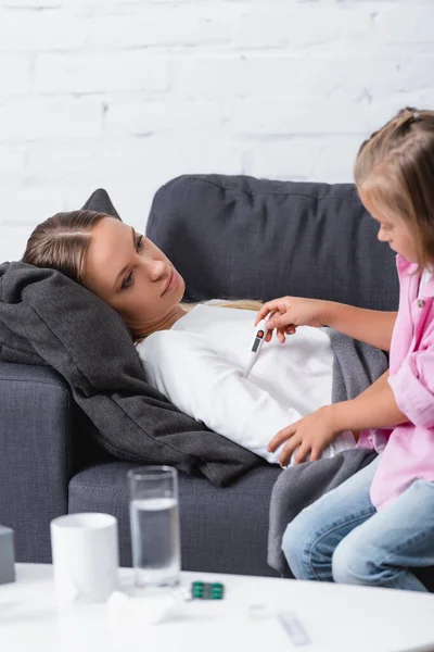 Selektiver Fokus des Kindes mit Thermometer in der Nähe einer kranken Frau in der Nähe von Tabletten und einem Glas Wasser auf dem Couchtisch — Stockfoto