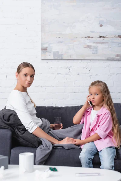 Selective focus of child talking on smartphone near sick mother with glass of water at home — Stock Photo