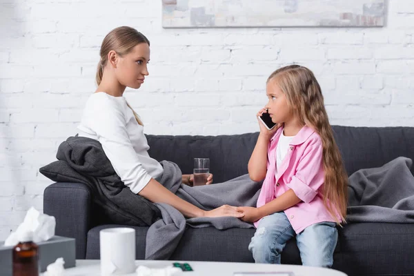 Selektiver Fokus des Kindes, das auf dem Smartphone spricht und die Hand der kranken Mutter mit einem Glas Wasser auf der Couch hält — Stockfoto