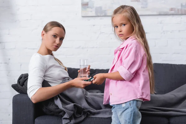Focus selettivo del bambino che guarda la fotocamera mentre dà pillole alla madre con un bicchiere d'acqua sul divano — Foto stock