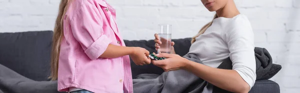 Панорамный снимок ребенка, дающего таблетки больной женщине со стаканом воды на диване — стоковое фото