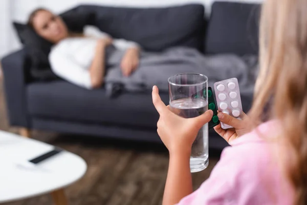 Enfoque selectivo del niño sosteniendo ampollas con pastillas y vaso de agua cerca de la madre enferma en casa - foto de stock