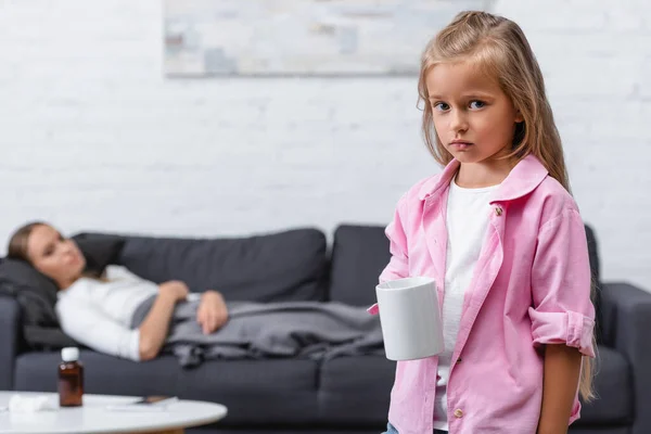 Selektiver Fokus eines traurigen Mädchens, das Tasse mit kranker Frau auf Couch im Hintergrund hält — Stockfoto