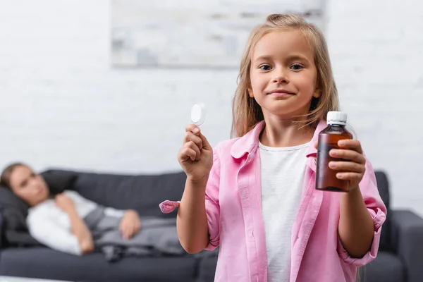 Enfoque selectivo de la niña sosteniendo cuchara y botella de jarabe cerca de la madre enferma en la sala de estar - foto de stock