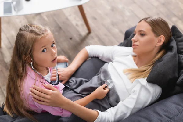 Vista ad alto angolo del bambino nello stetoscopio guardando la fotocamera vicino alla madre malata in soggiorno — Foto stock