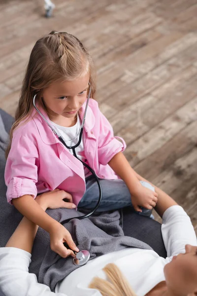 Vue grand angle de la femme malade couchée sur le canapé près de la fille avec stéthoscope — Photo de stock