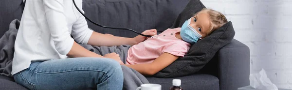 Horizontal image of child in medical mask looking at mother with stethoscope on couch — Stock Photo