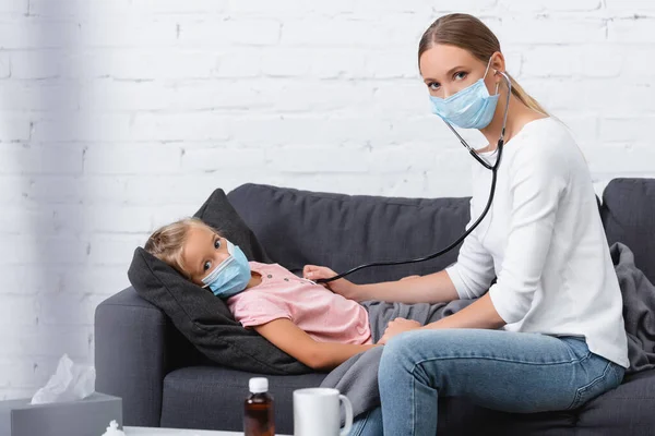 Selective focus of woman in medical mask using stethoscope near sick child on couch — Stock Photo