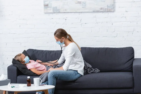 Selective focus of mother using stethoscope on sick daughter in medical mask — Stock Photo
