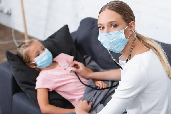 Focus selettivo della donna in maschera medica e stetoscopio seduto vicino al bambino a casa — Foto stock