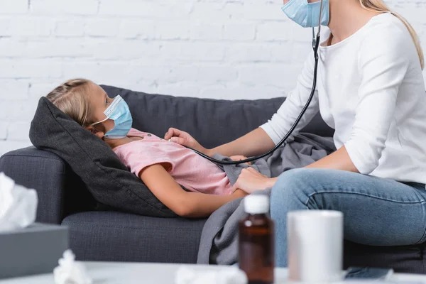 El foco selectivo de la mamá usando el estetoscopio a la hija en la máscara médica cerca de la taza y el jarabe en la mesa de café - foto de stock