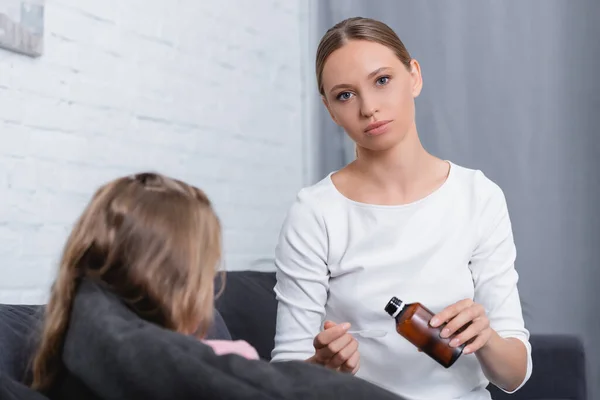 Selective focus of woman looking at camera while holding syrup and spoon near ill daughter on couch — Stock Photo