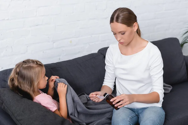 Bambino malato con coperta sdraiato vicino alla madre versando sciroppo sul divano — Foto stock