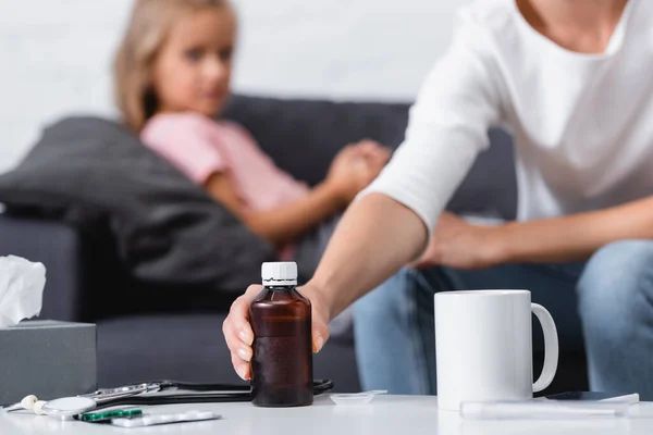 El foco selectivo de la mujer tomando el jarabe cerca de las pastillas y el estetoscopio en la mesa cerca de la hija enferma - foto de stock