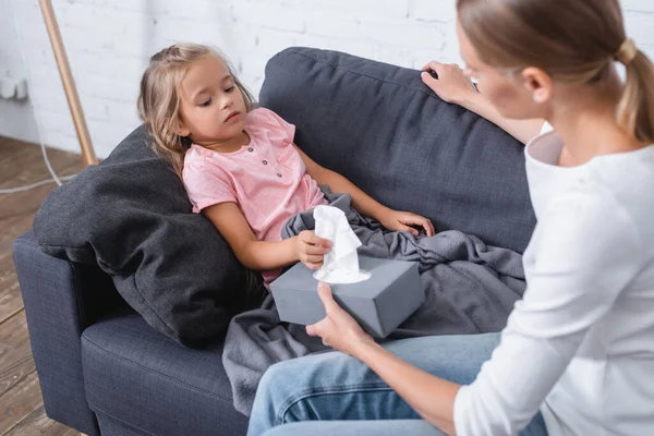 Enfoque selectivo de la niña que toma la servilleta cerca de la madre con la caja durante la enfermedad en el sofá en casa - foto de stock