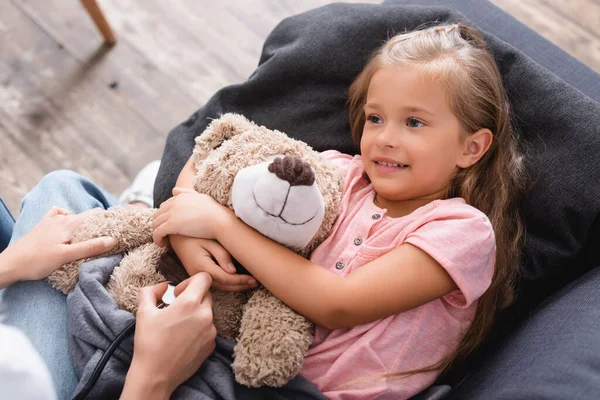 Enfoque selectivo de la mujer sosteniendo estetoscopio cerca de la hija abrazando oso de peluche en casa - foto de stock