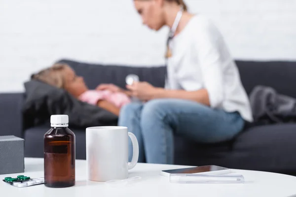 Foco seletivo de pílulas, xarope e smartphone na mesa perto da mãe sentada ao lado da criança doente em casa — Fotografia de Stock