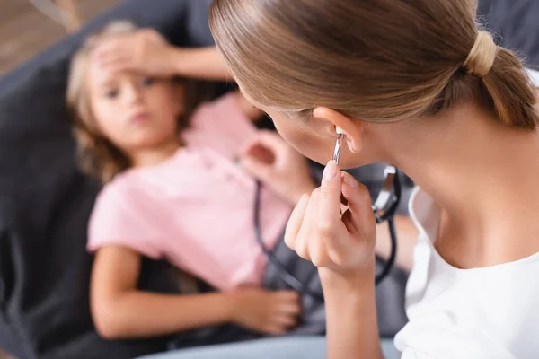 El foco selectivo de la madre en el estetoscopio sentado al lado de la hija enferma en el sofá - foto de stock