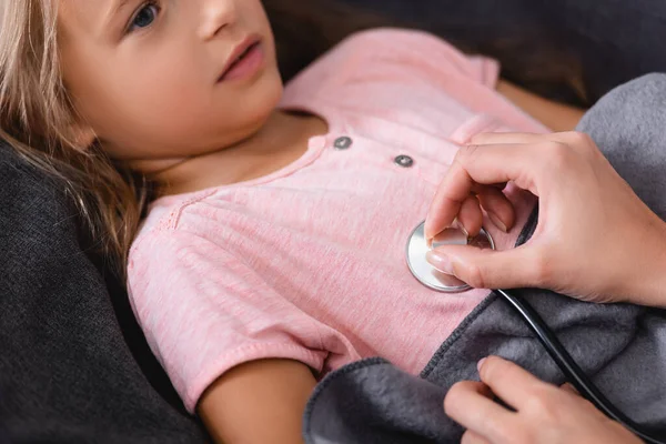 Vista cortada da mulher segurando estetoscópio perto da filha doente deitada no sofá — Fotografia de Stock