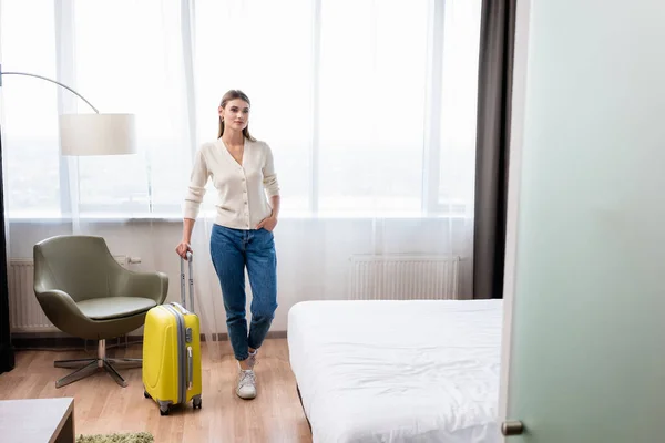 Молода жінка стоїть з рукою в кишені і жовтий багаж в готельному номері — стокове фото