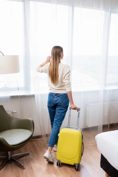 Vista trasera de la joven mujer de pie con equipaje amarillo en la habitación de hotel - foto de stock