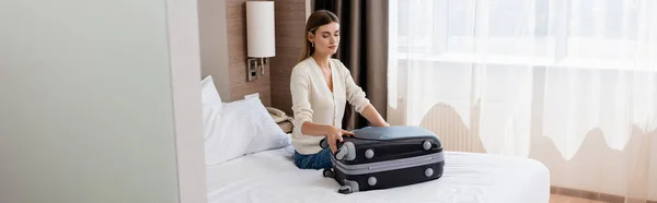 Imagen horizontal de la joven sentada en la cama y mirando el equipaje en la habitación de hotel - foto de stock