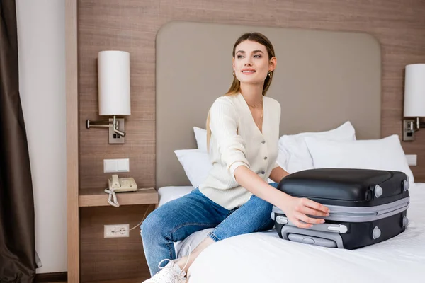 Mujer joven sentada en la cama y tocando el equipaje mientras mira hacia otro lado en la habitación del hotel - foto de stock
