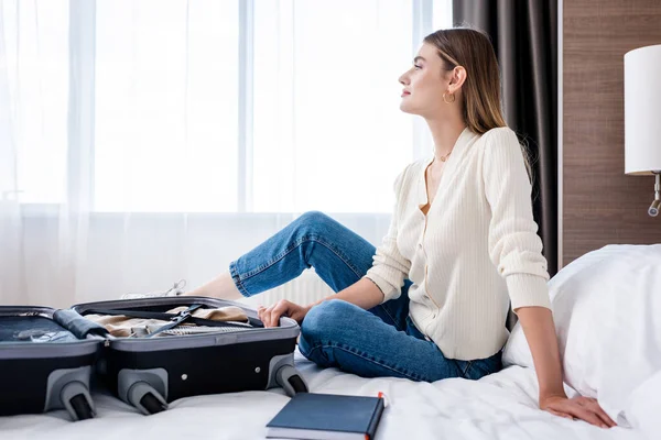 Vista lateral da jovem mulher sentada na cama e olhando para longe perto da bagagem no quarto do hotel — Fotografia de Stock