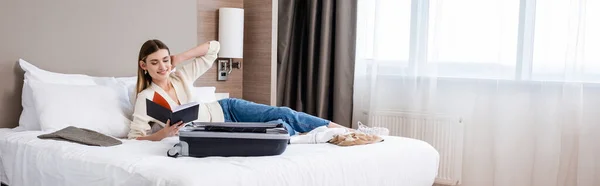 Panoramic concept of young woman looking at notebook near luggage on bed in hotel — Stock Photo