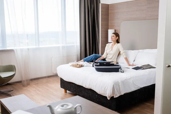 Селективный фокус молодой женщины, смотрящей в сторону возле ноутбука и багажа на кровати в отеле — стоковое фото