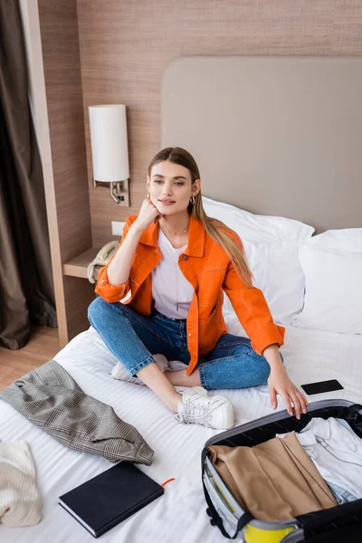 Joven viajero sentado en la cama cerca de la ropa, equipaje amarillo, teléfono inteligente y portátil en el hotel - foto de stock