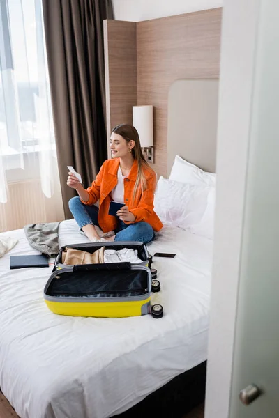 Woman holding passport, boarding pass and air ticket near baggage and notebook on bed in hotel room — Stock Photo