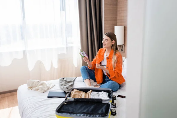 Selektiver Fokus der jungen Frau mit Pass, Bordkarte und Flugticket in der Nähe von Gepäck und Notizbuch im Hotelbett — Stockfoto