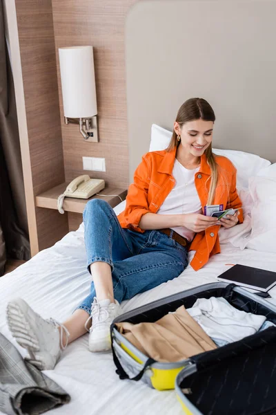 Gioiosa donna in possesso di passaporto, carta d'imbarco e biglietto aereo vicino al bagaglio e notebook sul letto in hotel — Foto stock