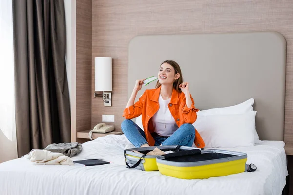 Viajante animado segurando passagem aérea perto de bagagem, roupas e notebook na cama no hotel — Fotografia de Stock
