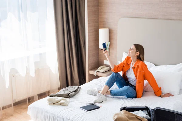 Zufriedene Frau beim Blick auf Reisepass und Flugticket in der Nähe von Gepäck und Kleidung auf Hotelbett — Stockfoto