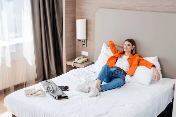 Молодая женщина отдыхает возле смартфона с чистым экраном и одеждой на кровати — стоковое фото