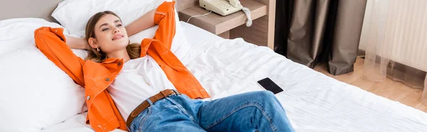 Панорамный снимок молодой женщины, отдыхающей возле смартфона с пустым экраном на кровати в отеле — стоковое фото