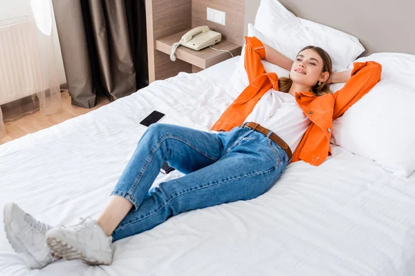 Mujer joven descansando cerca de teléfono inteligente con pantalla en blanco en la cama en el hotel - foto de stock