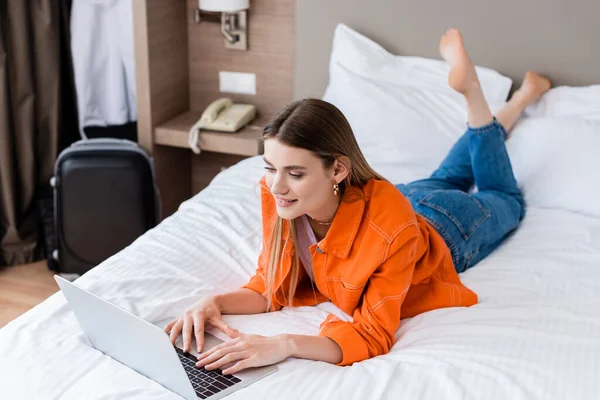 Barefoot freelancer lying on bed and using laptop in hotel room — Stock Photo