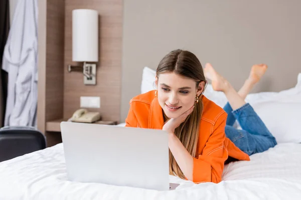 Jovem freelancer descalço deitado na cama e usando laptop no quarto de hotel — Fotografia de Stock