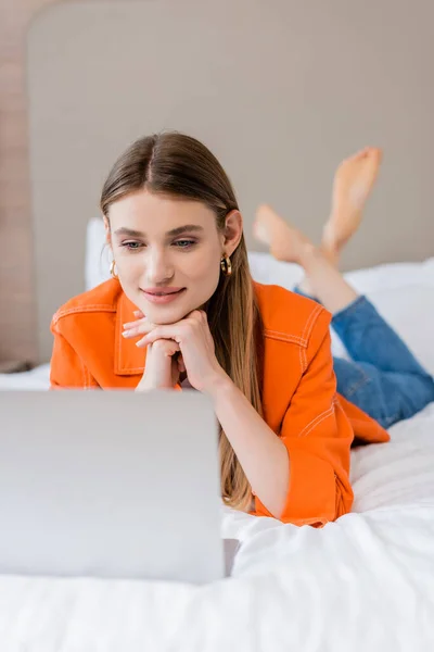 Jovem freelancer deitado na cama e usando laptop no quarto de hotel — Fotografia de Stock