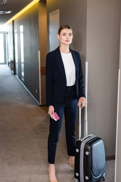 Businesswoman in suit holding room card while standing with luggage in hall of hotel — Stock Photo